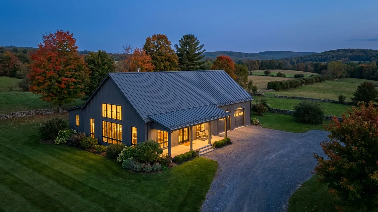 A finished barndominium at dusk among autumn maples, with warm interior lighting glowing through its large windows.