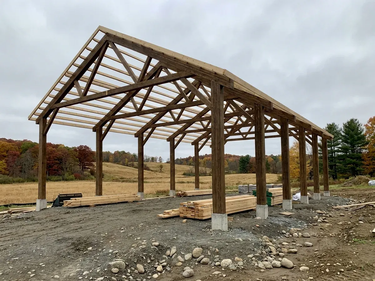 A pole barn home under construction, showing laminated timber columns on concrete piers and wide-span roof trusses with no interior bearing walls.