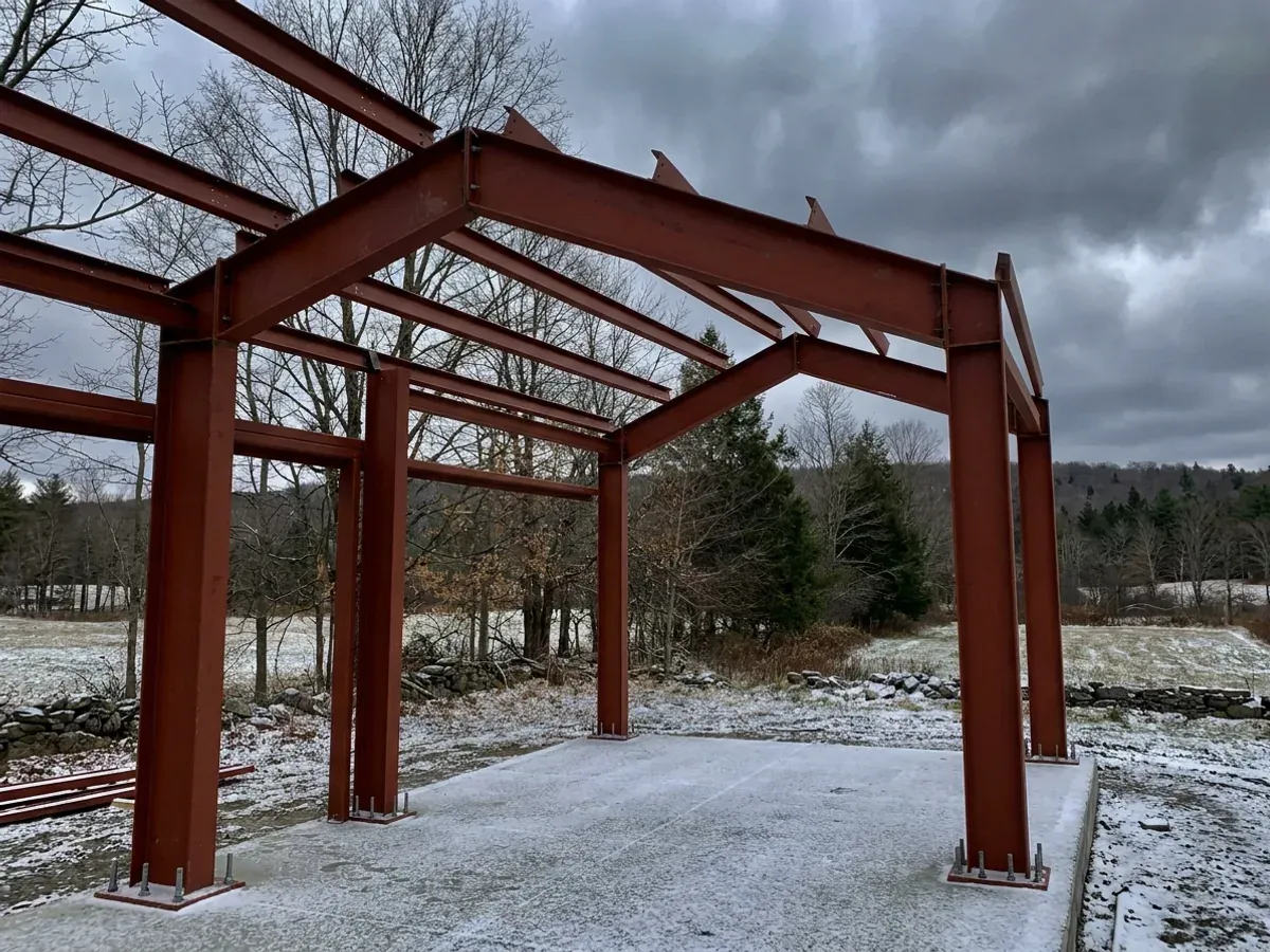 A red oxide primed structural steel frame partly erected on a concrete slab, showing bolted knee joints and anchor bolts.